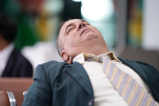A tired male passenger sleeps sitting up beside his luggage in an airport departure lounge, a familiar image of travel exhaustion, showing jet lag and long haul fatigue