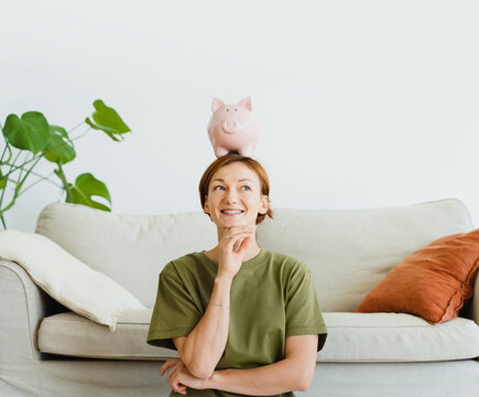 A smiling woman balances a pink piggy bank on her head indoors
