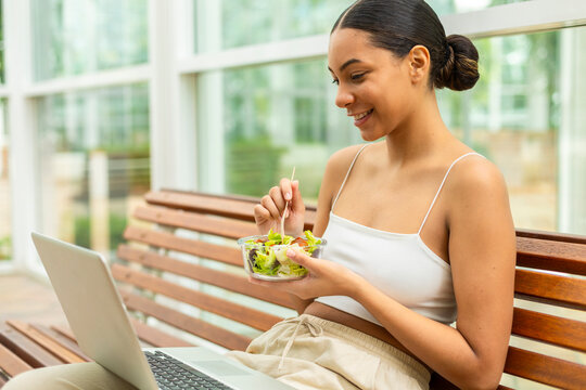 Young woman eats healthy salad while working on laptop in the park