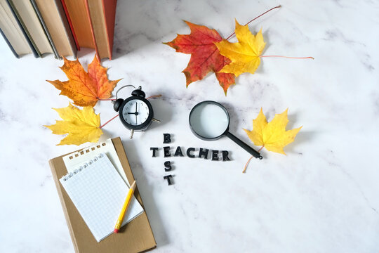 Teacher's day holiday, education concept. word "best teacher" made of wooden letters, school supplies, fallen maple leaves, loupe, alarm clock and books on table. greeting card design. flat lay