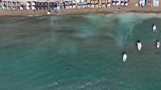 Aerial footage looking straight down over Laganas Beach on the Greek island of Zakynthos showing clear shallow sea swimmers small boats sun loungers and parasols lining the sandy shoreline