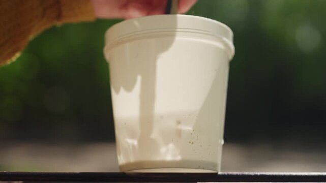 Close up shows hands stirring thick creamy off white acrylic paint in small round bucket. scene focuses on texture and consistency of mixed colors with soft natural light highlighting