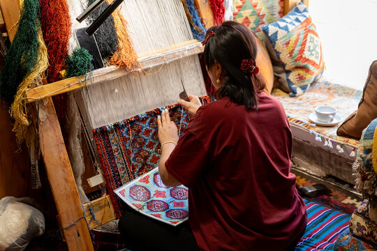 Bukhara, Uzbekistan. Rear view woman works at a wooden handloom, weaving a colorful patterned rug while textile samples, cushions, yarn, and tools surround her in warmly lit craft workshop interior