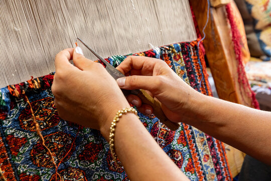Hands trimming colorful woven textile on traditional loom with scissors and patterned threads in a close up craft workshop scene. Woman works at a wooden handloom, weaving a colorful patterned rug