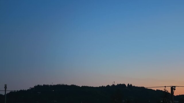 Subtle timelapse of a clear evening sky transitioning from dark blue to warm orange light at sunset above a dark treeline ridge silhouette