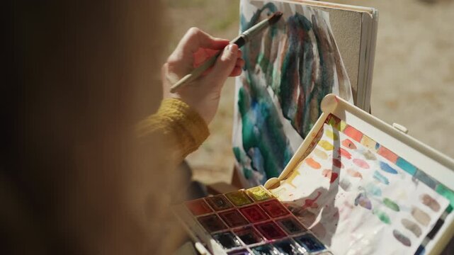 Close up shows person painting with watercolors outdoors. color palette and partially completed artwork are visible as hand dips brush into paint and applies it to canvas focusing