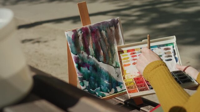Person wearing yellow sleeves mixes colors with brushes from tray of paints then applies them onto partially completed watercolor painting on easel. scene is set outdoors in natural light