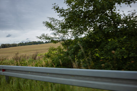 Country Road Guardrail Beside Agricultural Field and Trees. Rural roadside scene photographed from a moving vehicle, showing a silver metal safety guardrail in the foreground beside dense green vegeta