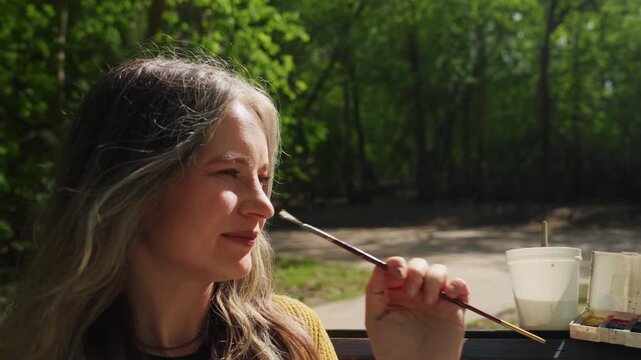 Woman enjoys painting outdoors holding brush by her head. white container of paints sits on bench beside her with green trees and path visible in background