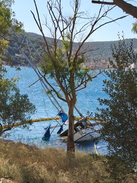 Partially sunken sailboat near the coast of Ithaca island, Greece, surrounded by a yellow oil containment boom. The image captures an environmental response scene, showing pollution prevention efforts