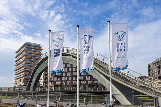 Purmerend, Netherlands - 27 September 2026: Modern Melkwegbrug pedestrian bridge with waving city flags and contemporary residential architecture against a cloudy blue sky.