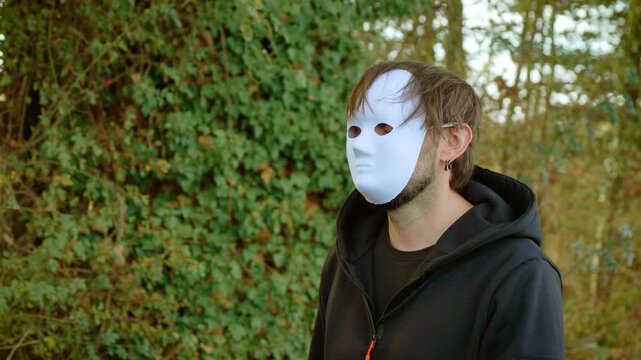 Masked killer walking through woodland in black hoodie with white disguise. Criminal murderer and maniac concept among trees and bushes. Violent crime, rapist, victim and mentally ill offender theme