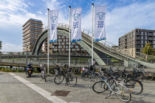 Purmerend, Netherlands - 27 September 2026: Group of parked bicycles in front of the modern Melkwegbrug bridge, city flags, and contemporary apartment buildings on a sunny day.