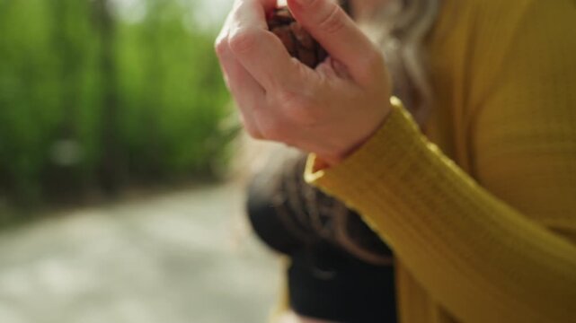 Woman with long hair wearing yellow cardigan examines small objects including handling natural materials like cones. scene is set outdoors with greenery in background