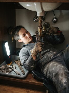A woman uses a pipe wrench to repair a leaking pipe beneath a sink, working in a dimly lit space illuminated by a portable work light.