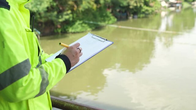 Water management engineer writes inspection notes on clipboard beside canal while checking water conditions and documenting findings during field survey.