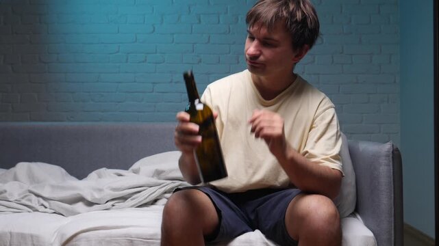 Young man sitting on a bed in a dark room, deeply immersed in depression and alcoholism, drinking from a bottle, representing a problem of substance abuse and loneliness