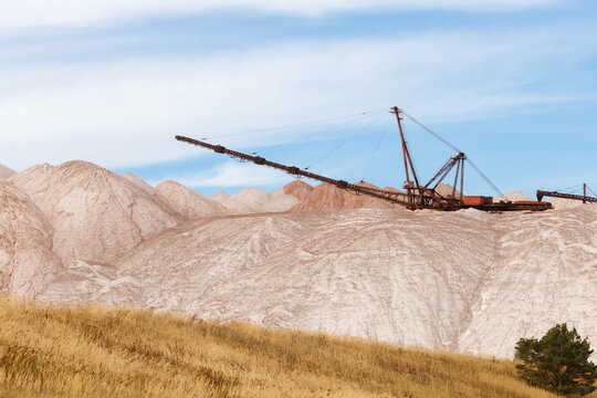 Large industrial spreader machine working on top of white and pink salt heaps or mining waste piles at a potash quarry under a blue cloudy sky horizontal view