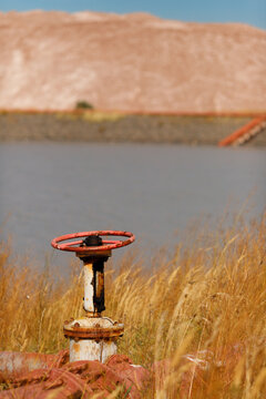 Vertical shot of an old rusty industrial metal pipe valve with a red handwheel wheel valve in a dry grass field near a technical water reservoir pond and massive brown salt slag heaps