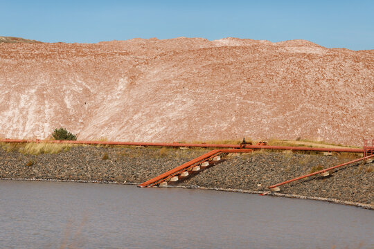 Industrial pipeline system installed on a gravel embankment of a technical reservoir pond near massive brown salt slag heaps at a potash mining quarry daytime horizontal view