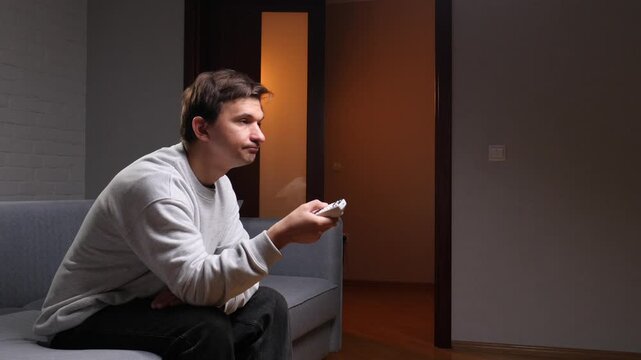 Man holding a remote control and watching television while sitting on a comfortable sofa in a dimly lit room during the evening, enjoying home entertainment