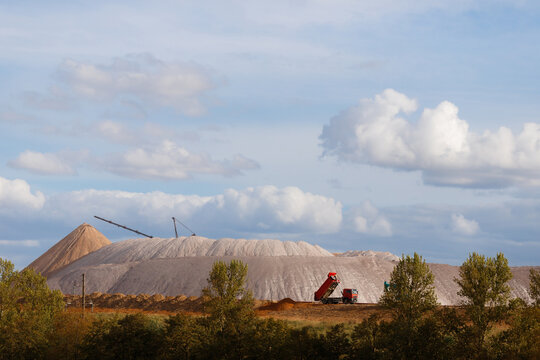 Red dump truck and blue excavator working on a soil embankment with massive white salt slag heaps on the background under a cloudy sky horizontal landscape view