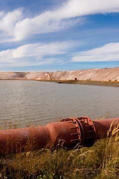 Vertical shot of a large industrial rusty iron pipeline on the grass shore of a technical water reservoir pond with massive brown salt slag heaps under a blue cloudy sky