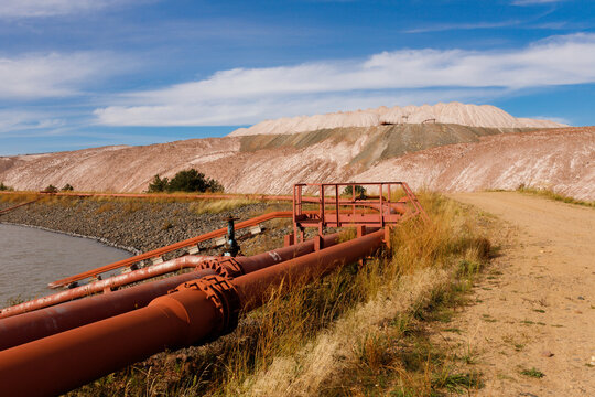 Industrial pipeline system with metal pipes and valve infrastructure running along a dirt road near technical water reservoir pond and massive brown salt slag heaps daytime horizontal landscape view