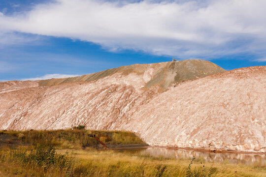 Massive multi layered brown and white salt slag heaps of a potash mining quarry rising behind a technical water reservoir pond under a blue sky with clouds daytime wide horizontal landscape view