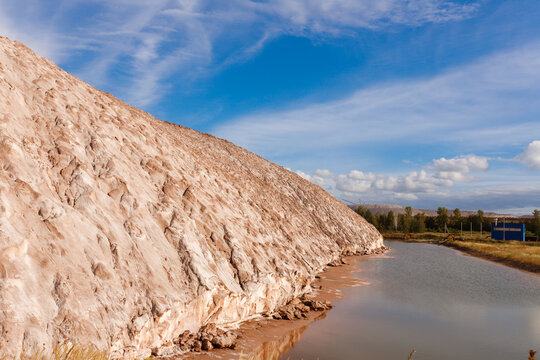 Close up view of a massive patterned brown and white salt slag heap slope next to a technical water reservoir pond of a potash mining quarry under a blue cloudy sky horizontal landscape shot