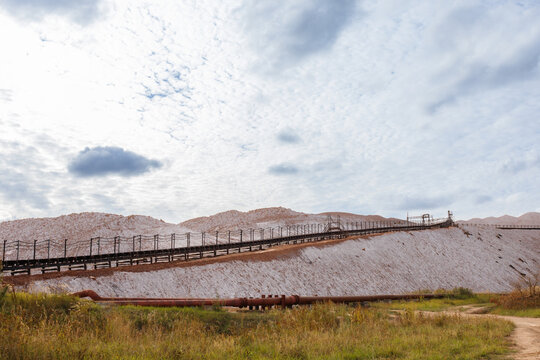 Long industrial steel conveyor belt gallery installation built on a slope of massive grey salt slag heaps of a potash mining quarry under a cloudy sky daytime wide horizontal landscape view