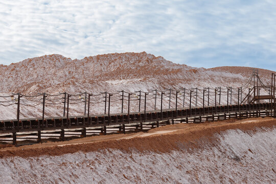 Long industrial steel conveyor belt gallery installation built on a slope of massive grey salt slag heaps of a potash mining quarry under a cloudy sky daytime wide horizontal landscape view