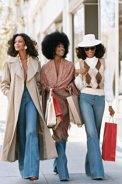 A group of three women carrying shopping bags walk along a city sidewalk.