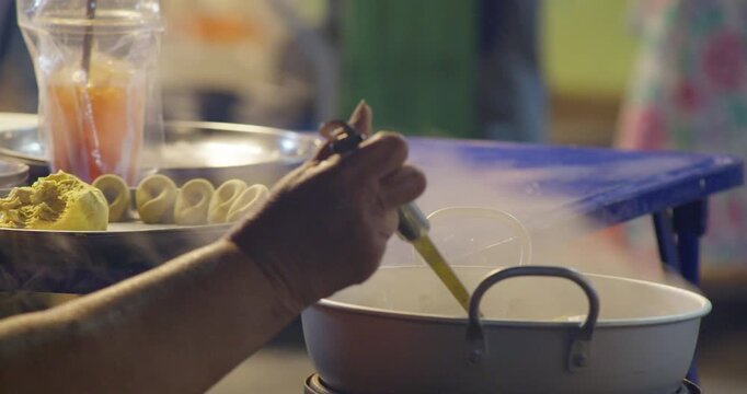 A street food vendor stirs rice flour dumpling balls boiling in a pot, with a plate of raw dough nearby at a Thai dessert stall.
