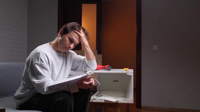 Frustrated man sitting on sofa, reading paper instructions and struggling to assemble flat pack furniture at home, concentrated and confused during diy project in living room
