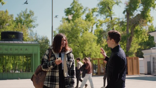 Young woman stands outdoors holding white phone as people walk around and many pigeons fly above. Woman wears plaid jacket with headphones carrying brown bag while another woman approaches