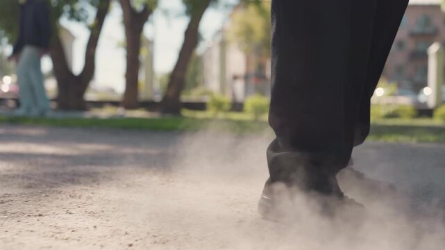 Focus remains fixed low to ground capturing pair of legs and feet as they walk on dusty pavement. Each footfall stirs up small clouds of dust emphasizing texture detail in motion. Background