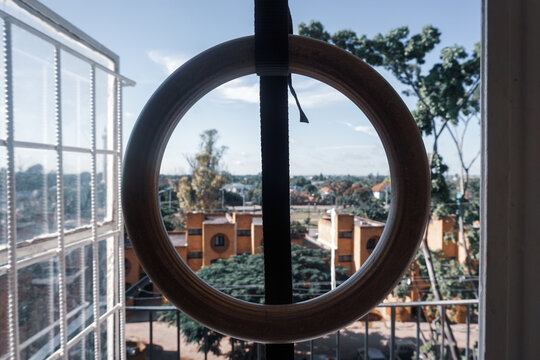 A wooden gymnastic ring hangs near an open window, framing a sunlit view of city buildings and lush green trees under a bright blue sky.