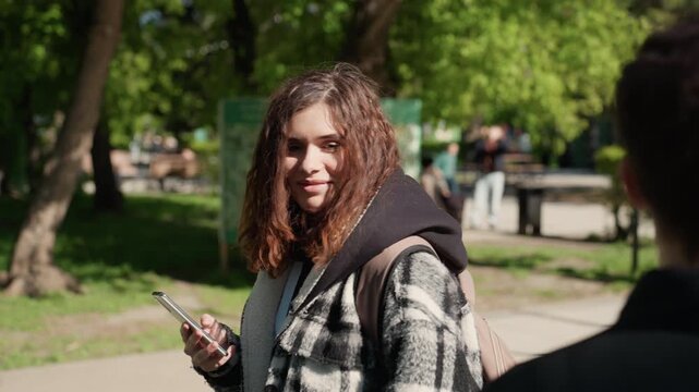 Young woman stands outdoors holding mobile phone while facing away from camera in park setting with another woman partially visible. Woman turns to face forward suggesting interaction