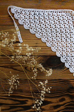 Delicate white handmade crochet lace kerchief scarf paired with gentle dried gypsophila flowers on a dark rustic wooden background. Romantic boho vintage fashion and lifestyle flat lay.