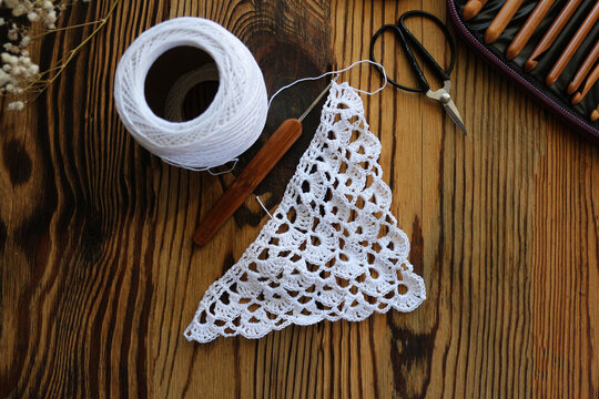 Unfinished white crochet lace piece with bamboo hook, yarn clew, scissors, and hook case on a dark rustic wooden table. Top view craft concept for handmade, DIY, and needlework projects.