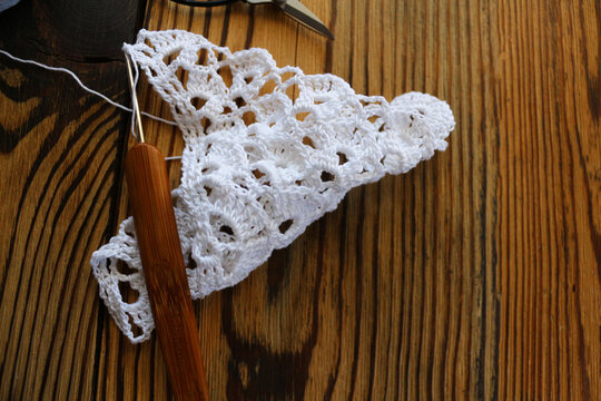 Close up of a white handmade crocheted lace piece with a wooden crochet hook on a dark rustic wooden background. Creative handcraft, needlework, DIY, and vintage hobby concept photography.
