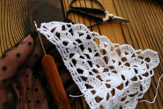 Close up of a female hand in elegant black polka dot glove holding wooden hook and crocheting white lace. Rustic wooden table with vintage scissors. Creative needlework and DIY concept.