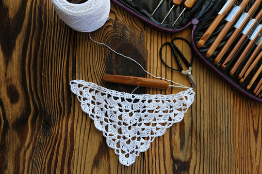 Unfinished white crochet lace piece with bamboo hook, yarn clew, scissors, and hook case on a dark rustic wooden table. Top view craft concept for handmade, DIY, and needlework projects.