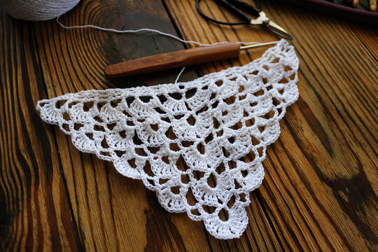 Close up of a white handmade crocheted lace piece with a wooden crochet hook on a dark rustic wooden background. Creative handcraft, needlework, DIY, and vintage hobby concept photography.