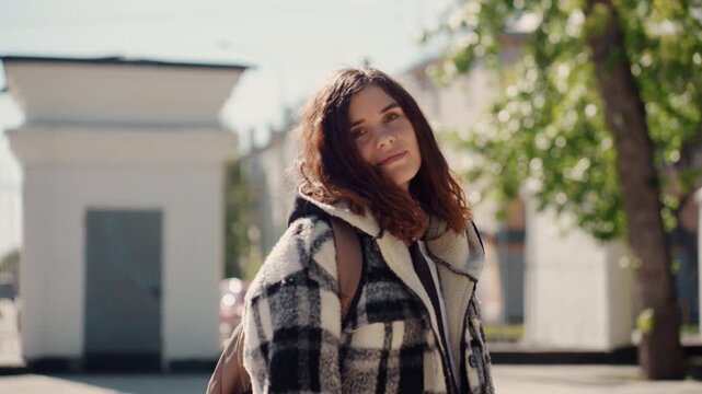 Woman stands on side of city streets. Woman wears casual clothing including headphones around her neck and carries bag. background shows buildings and greenery