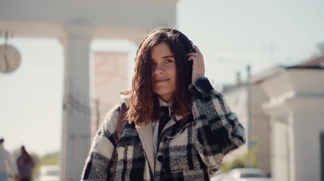 Young woman stands outside during day. Woman wears black and white checkered coat and large over ear headphones while holding them set against blurred urban street scene with buildings
