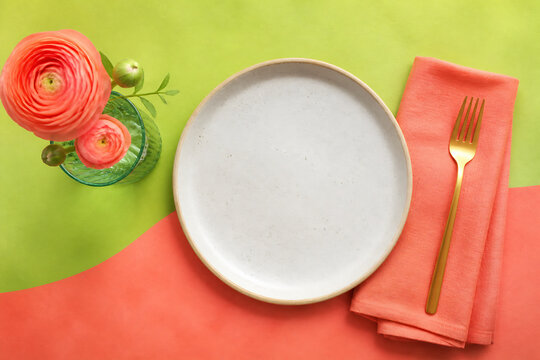 A white plate, a coral napkin with a gold fork, and pink flowers in a glass are arranged on a green and orange surface.