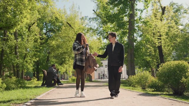 Woman and man walk together in park. background features green trees with sunlight filtering through leaves creating bright natural light. person sits on bench to left