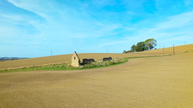 Abandoned stone farmhouse ruins under blue sky. Weathered gable wall and low masonry structures beside harvested fields. Rural countryside architecture among rolling farmland with utility poles and
